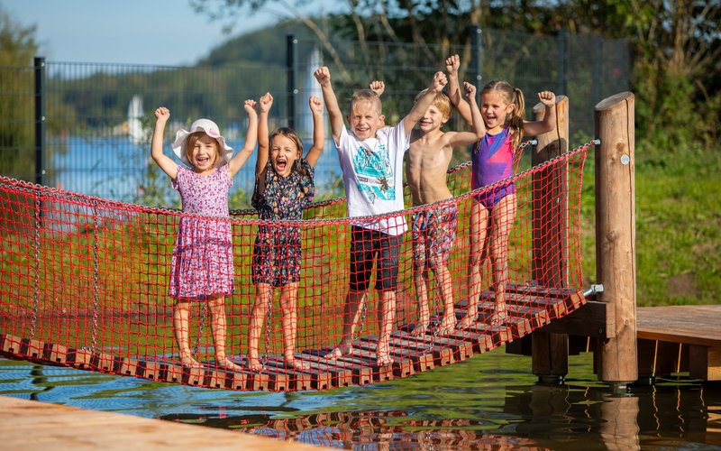 Wasserspielplatz Fünf Kinder stehen auf einer Hängeseilbrücke und strecken die Arme in die Luft.