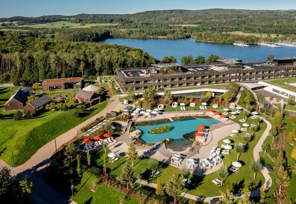 In einer Luftaufnahme des Wellnesshotels Seezeitlodge ist das Gebäude des Hotels, das keltische Saunadrof sowie der Außenschimmbereich mit Sonnenliegen und Bar zu sehen. Die Umgebung ist in eine grüne Landschaft eingebettet.