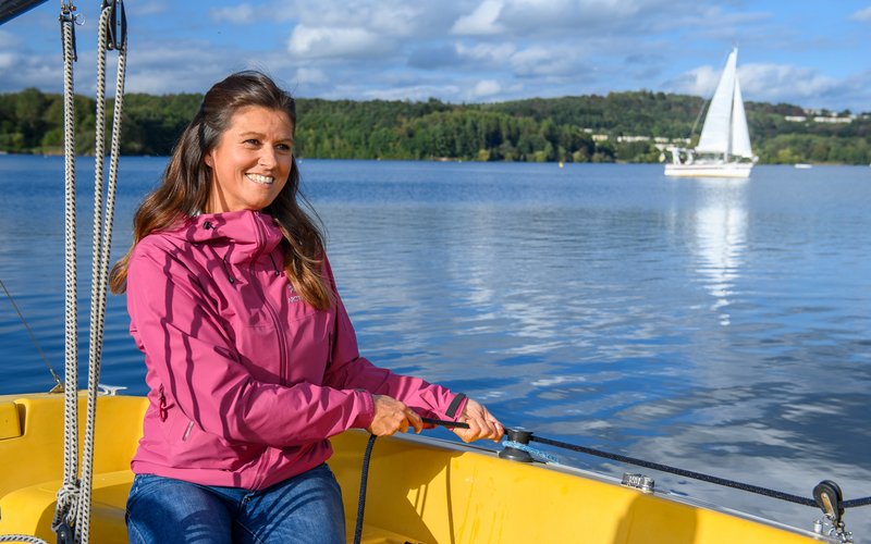 Wassersportmöglichkeiten am Bostalsee Eine junge Frau in Outddorkleidung sitzt auf dem gelben Segelboot der S.A.L.T Yacht GmbH und zieht die Segel an. Ihr Blick ist freundlich, in Richtung Kamera gerichtet. Hinter ihr sind das blaue Wasser des Bostalsee und ein Segelboot zu sehen.