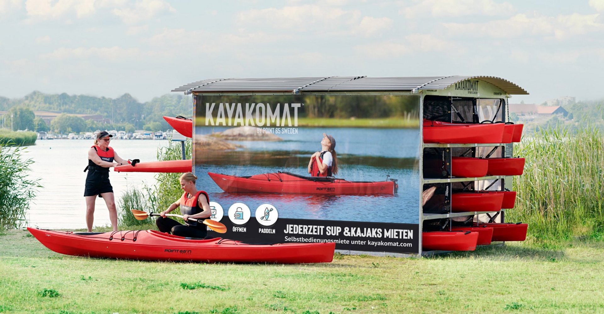 Eine Kajak Verleihstation an einem See. In einem überdachten Gehäuse sind mehrere Kajaks sowie SUP-Boards gelagert. Eine Frau ist vor einem auf dem Boden liegenden Kajak in der Hocke und baut ein Paddel zusammen und eine andere Frau zieht ein Kajak aus der Verleihstation.