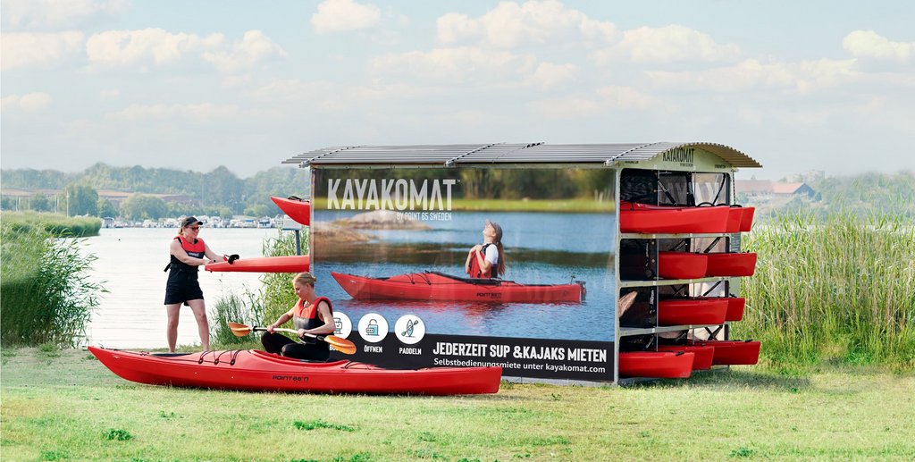 Photo: Malte Danielsson AB Eine Kajak Verleihstation an einem See. In einem überdachten Gehäuse sind mehrere Kajaks sowie SUP-Boards gelagert. Eine Frau ist vor einem auf dem Boden liegenden Kajak in der Hocke und baut ein Paddel zusammen und eine andere Frau zieht ein Kajak aus der Verleihstation.