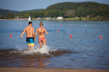 Ein junges Paar ist von hinten zu sehen, während es Hand in Hand in den Badesee Bostalsee läuft.