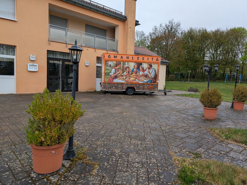 Foodtruck Mahlzeit Frontalsicht auf einen geschlossenen Foodtruck, auf dem ein Gemälde abgebildet ist. Um den Foodtruck befinden sich Pflanzenkübel, die zu einer gemütlichen Atmosphäre beitragen.