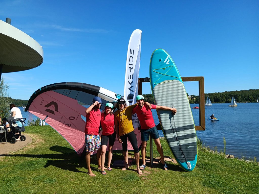 Foto: Lakeride 4 Personen stehen am Ufer des Bostalsees und haben ein Wingfoil, ein SUP und eine Beachflag mit dabei.