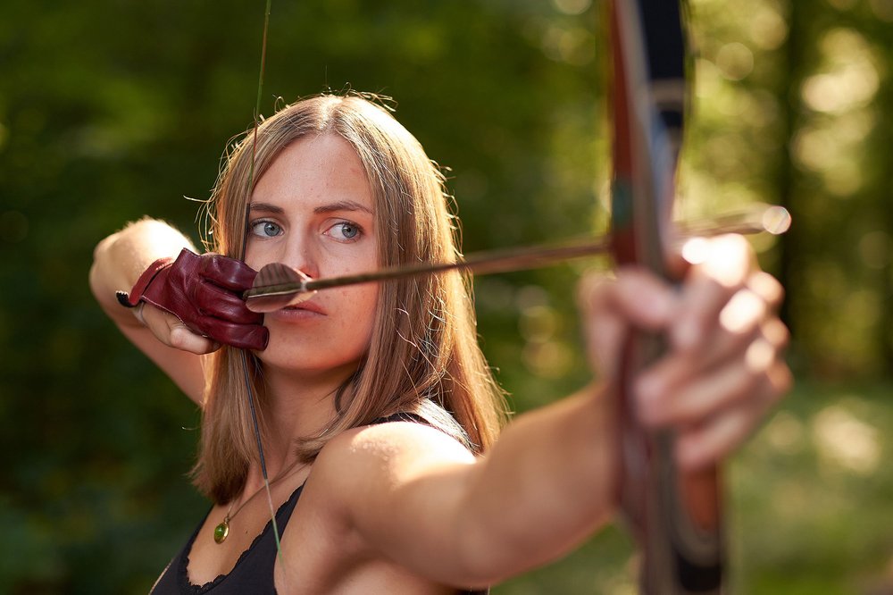 Das Bild zeigt eine junge, blonde Frau mit Pfeil und Bogen aus der Nahaufnahme. Sie schaut konzentriert auf ihr anvisiertes Ziel.
