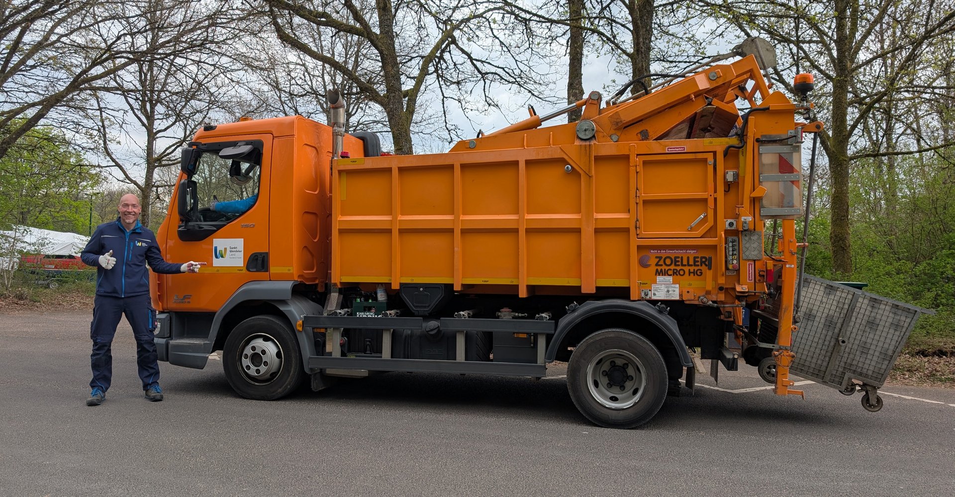 Ein orangenes Müllfahrzeug mit einem der Bauhofmitarbeiter, die für das Sammeln des Mülls zuständig sind, im Vordergrund. Er zeigt mit einer Hand auf das Fahrzeug und hält den Daumen hoch.