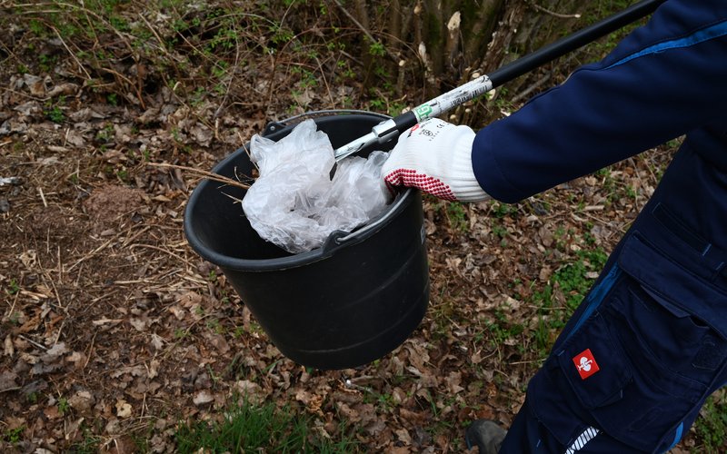 Eine Person hält einen schwarzen Eimer in ihrer behandschuhten Hand. In der anderen Hand hält sie eine Müllzange, mit der sie eine Plastiktüte in den Eimer fallen lässt.