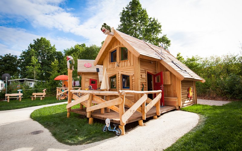 Im Fokus des Fotos befindet sich ein Märchenhaus. Es ist ein kleines Holzhaus, mit einer Terrasse und Spielgerät.