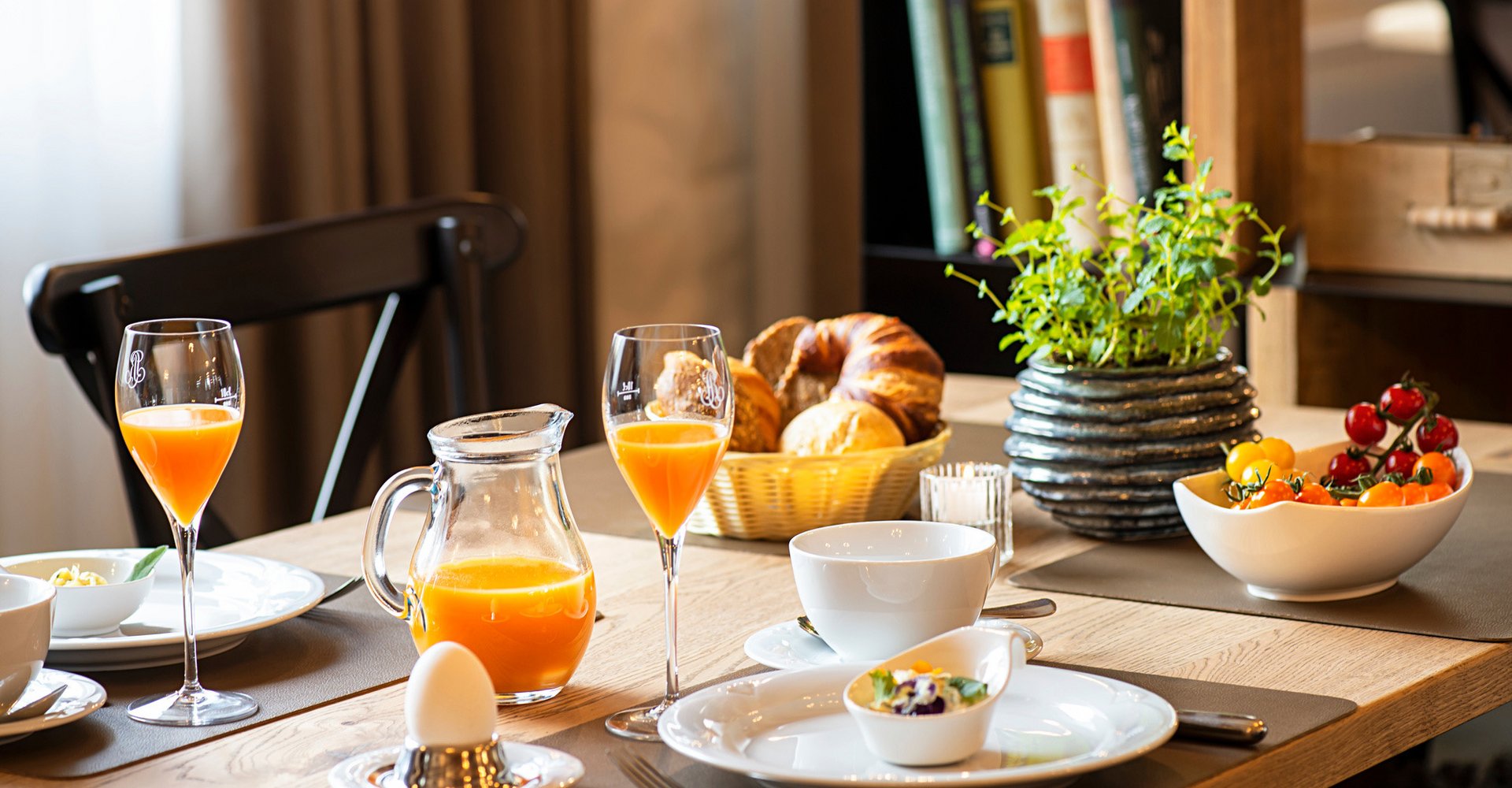 Auf dem Frühstückstisch im Victor's Seehotel Weingärtner stehen ein Brotkorb mit Brötchen und Croissants, Orangensaft, Frühstückseier und eine Schüssel mit Tomaten. 