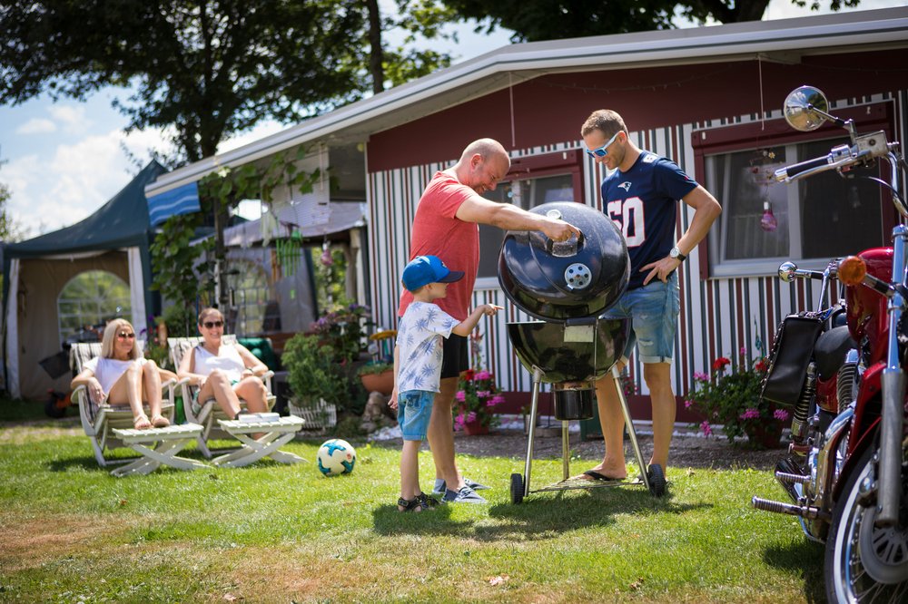 Familienzeit auf dem Campingplatz Zwei Männer und ein Junge stehen an einem Kugelgrill vor einem Wohnwagen mit Vorzelt, während zwei Frauen in Liegestühlen liegen und den Männern zusehen.