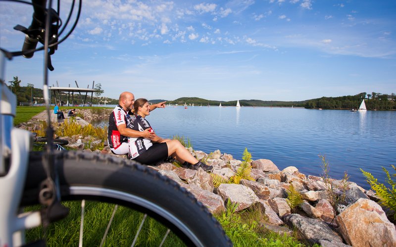 Ein Paar in Radsportkleidung sitzt im Gras am Seeufer des Bostalsees mit Blick aufs Wasser. Der Herr zeigt auf etwas in der Ferne mit ausgestrecktem Arm und Finger. Der vordere Teil eines Fahrrads ist links im Vordergrund des Bilds zu sehen.