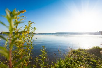 Vom Ufer aus hat man einen tollen Blick auf das blaue Wasser des Bostalsees mit der Sonne im Hintergrund.
