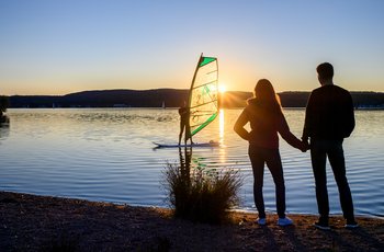Wassersportmöglichkeiten am Bostalsee bei Sonnenuntergang auf dem Bostalsee steht ein Windsurfer auf seinem Bord und schaut zum Ufer, wo gleichzeitig ein junges Pärchen Hand in Hand steht.