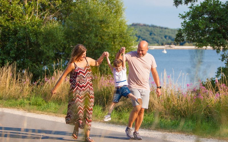 Ein Paar mit einem kleinem Jungen geht auf dem Seerundweg spazieren. Sie spielen mit dem Jungen das Spiel "Engelchen flieg". Er wird von beiden an seinen Armen in die Luft gehoben. Im Hintergrund ist ein Blick auf den See zu erhaschen.