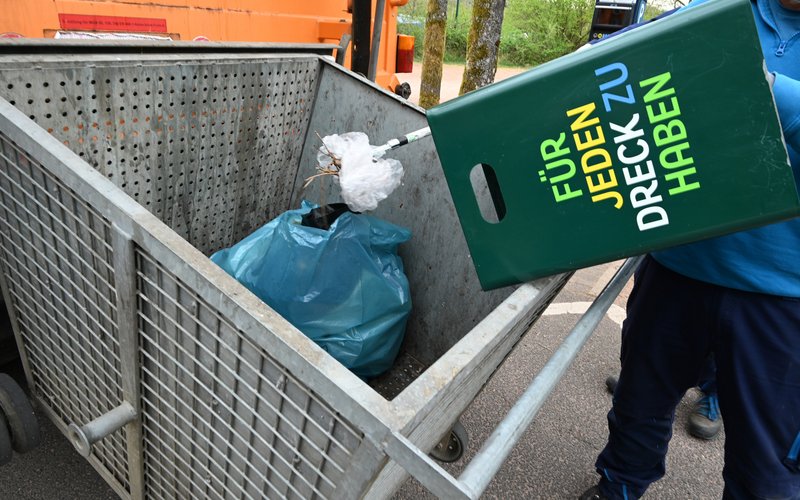 Ein Mann leert einen grünen Mülleimer mit der Aufschrift "Für jeden Dreck zu haben" in den Fangkorb eines Müllwagens.