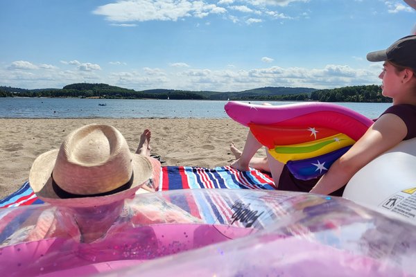 Eine junge Frau und ein junger Mann verweilen im Sand des Strandbad Bosens. Jeweils angelehnt an ein Schwimmtier, die Frau an einem Einhorn, der Herr an einem Famingo, liegen sie auf einer bunten Decke und genießen das gute Wetter und richten ihr Blick aufs Wasser.