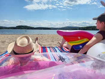 Eine junge Frau und ein junger Mann verweilen im Sand des Strandbad Bosens. Jeweils angelehnt an ein Schwimmtier, die Frau an einem Einhorn, der Herr an einem Famingo, liegen sie auf einer bunten Decke und genießen das gute Wetter und richten ihr Blick aufs Wasser.
