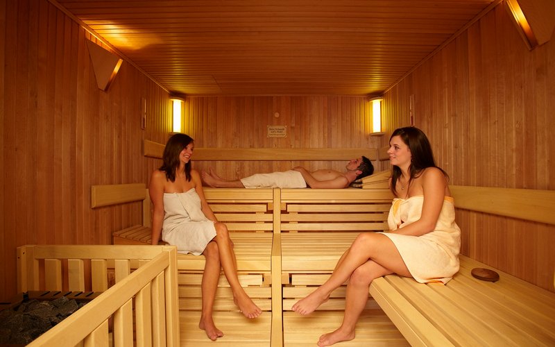 In einer aus Holz gebauten Sauna halten sich zwei Frauen und ein Mann auf. die Frauen unterhalten sich im Sitzen, währned der Herr im Hintergrund im Liegen entspannt.