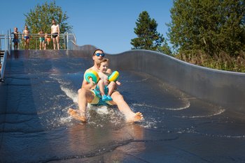 Ein junger Mann, rutsch mit Kleinkind in Schwimmflügeln eine wellenförmige Wasserrtusche herunter. Oben, am Rand der Rutsche warten weitere Personen, um ebenfalls herunterrutschen zu können.