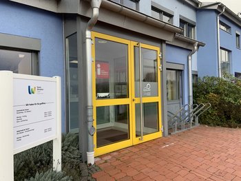 Die gelbe Eingangstür der Tourist-Information ist geschlossen. Daneben steht eine Tafel mit den Öffnungszeiten.