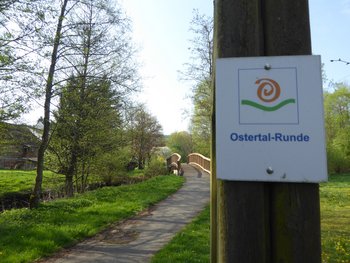 Ein asphaltierter Weg führt über eine Brücke zu einem Gebäude. An einem Pfosten vorne rechts hängt ein Wegweiser mit der Aufschrift "Ostertal-Runde".
